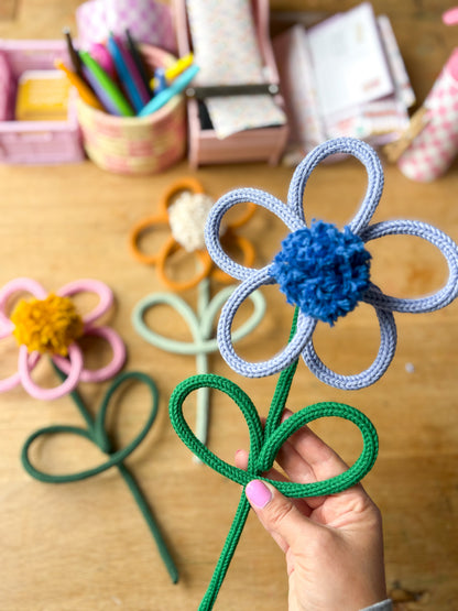 A hand holds a handmade rope/yarn flower with a blue pom-pom center and light blue looped petals, with other colorful rope flowers and a wooden desk in the background.
