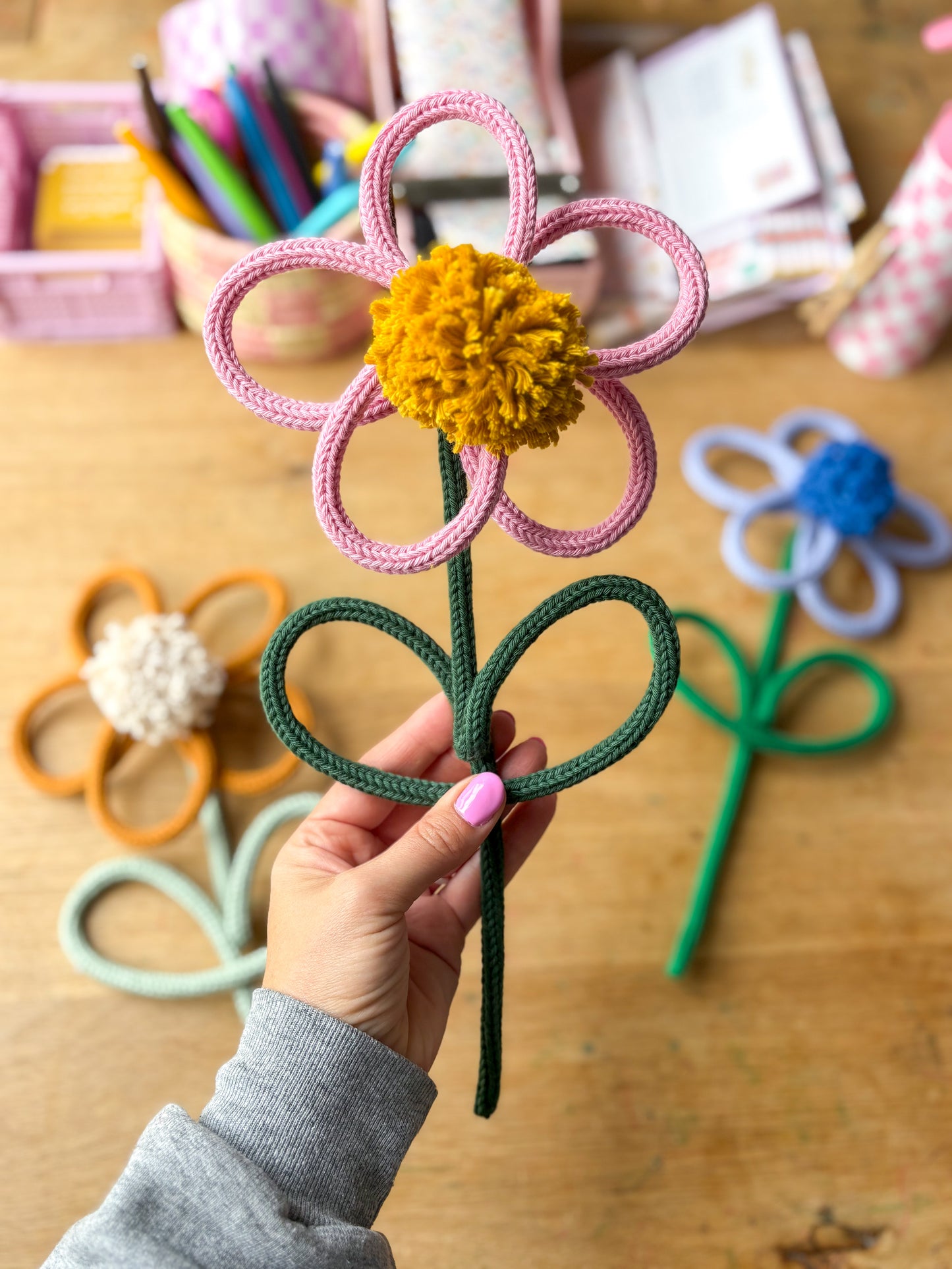 A hand holds a handmade rope - yarn flower craft with a pink circular bloom, yellow pom-pom center, and dark green stem on a wooden desk with other colorful rope flowers in the background.