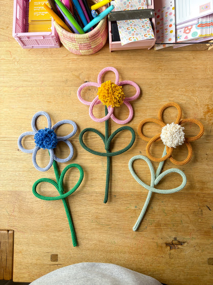 Three knotted rope flowers in pink, orange, and blue-gray are arranged on a wooden desk, with colorful stationery baskets visible in the background.