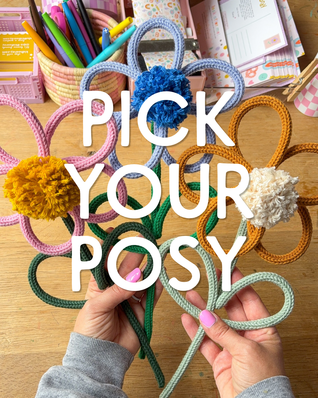 A person's hands hold several colorful rope flower accessories with pom-poms on a wooden desk, with large white text overlay reading 'PICK YOUR POSY'.