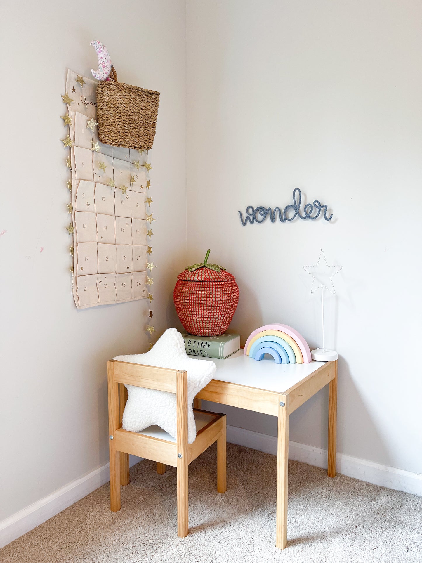 Knitted wire word "wonder" hanging on wall above kid's desk. The desk has a stacked wooden rainbow, strawberry basket. It features muted colors and lots of natural wood.