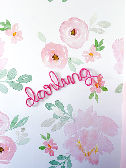 Knitted wire word "darling" in pink hanging on nursery wall with floral print wallpaper.