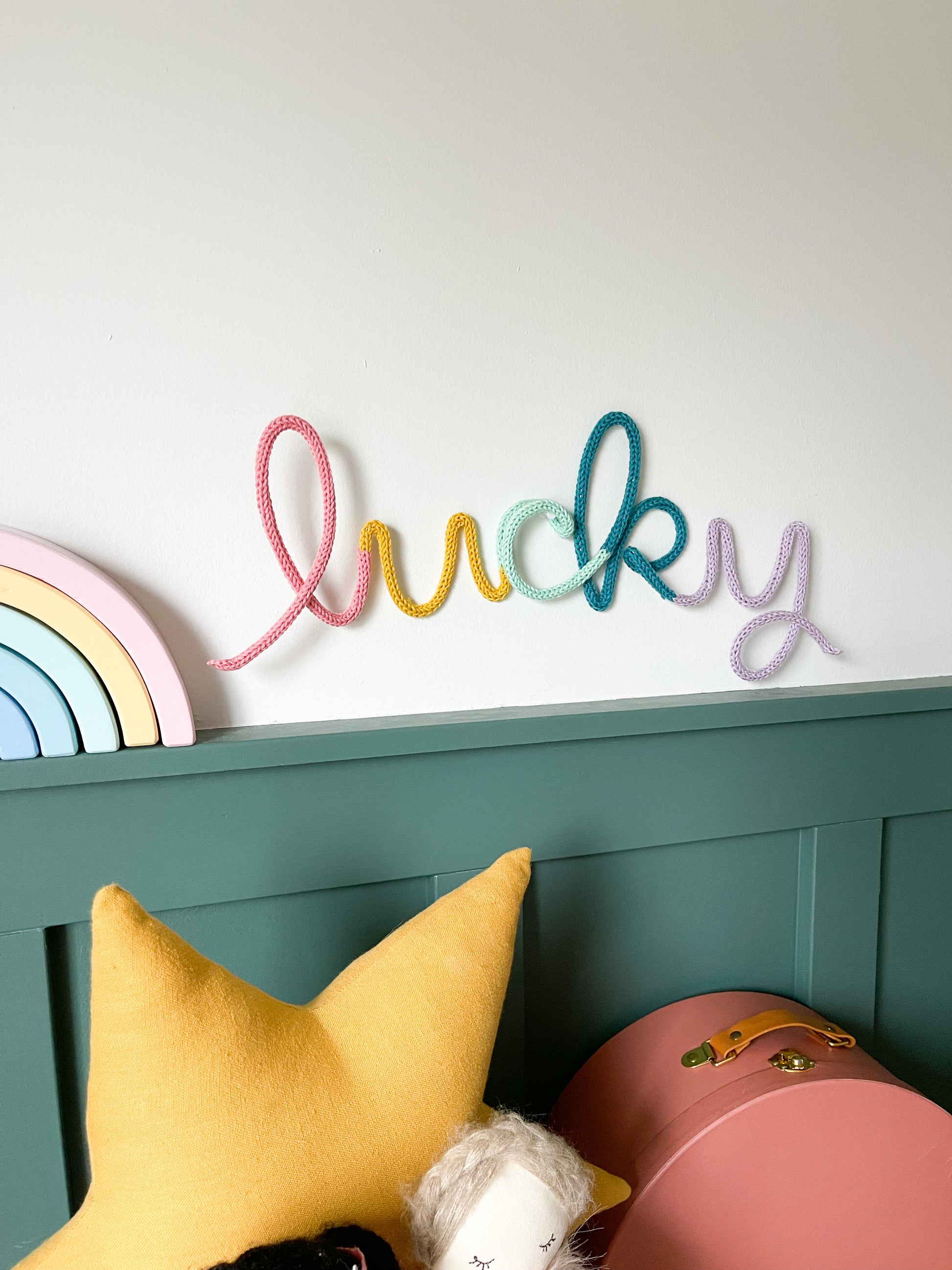 Knitted wire word lucky in rainbow color scheme. It's hung above a child's desk. 