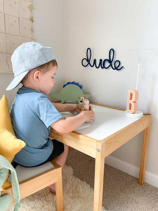 Knitted wire word "dude" sign. It's hung above a desk in a little boy's room. The boy is sitting at the desk playing with toys.