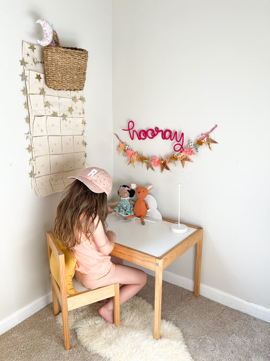 Knitted wire word "hooray". It's hung above a desk in a corner of a girl's bedroom. The little girl is sitting and coloring at her desk. 