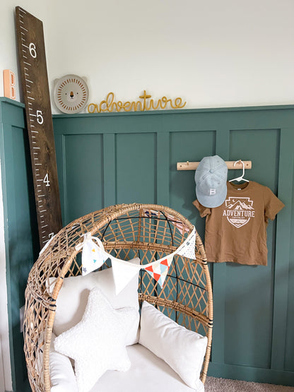 Knitted wire word "adventure" in a mustard yellow color. It's propped up on a ledge in a boys nature themed bedroom with hunter green board and batten.
