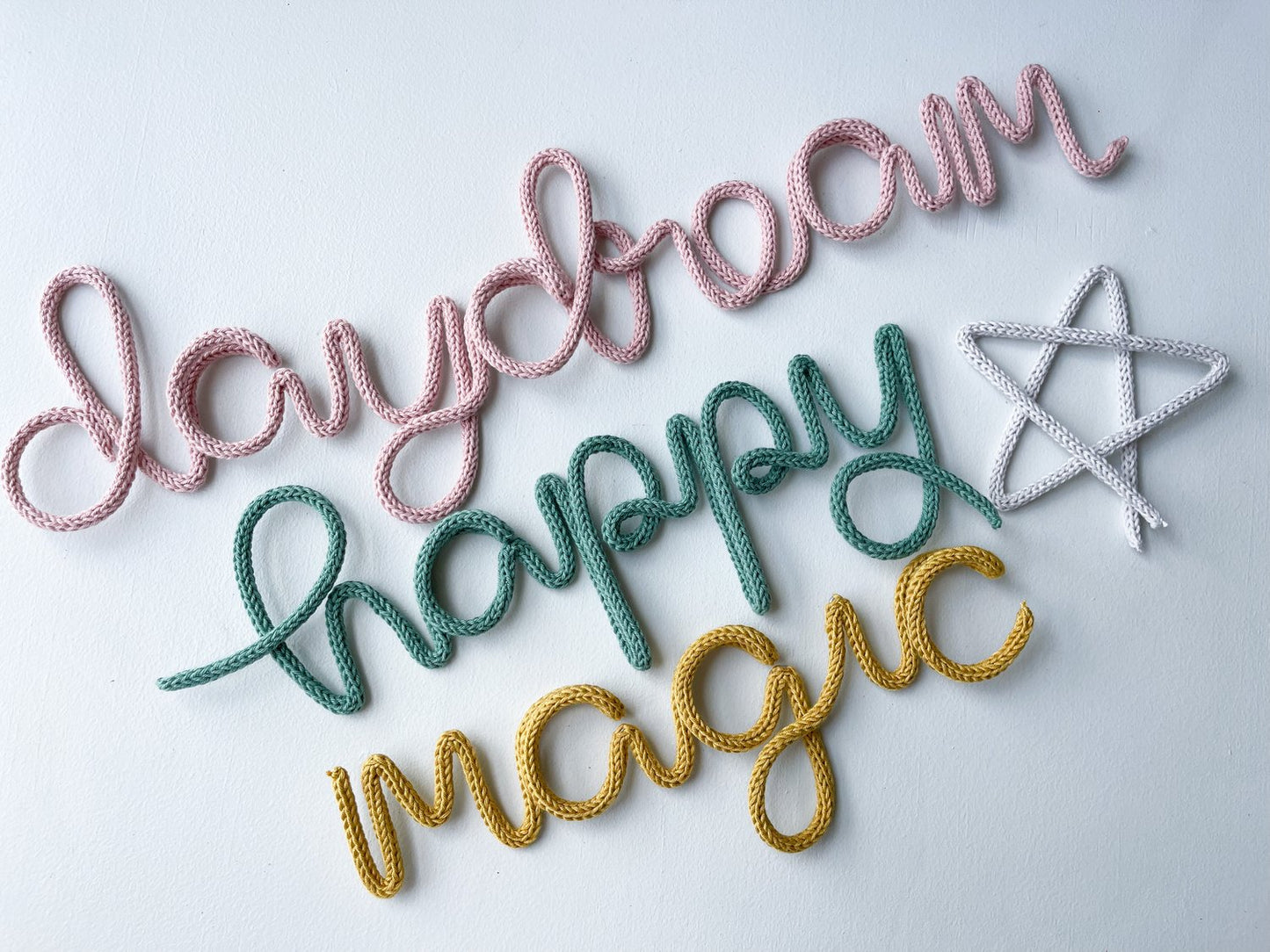 Flat lay style photo feature knitted wire words and wire art: daydream in the color blush, happy in the color sage green, magic in mustard and a whimsical star in light gray.