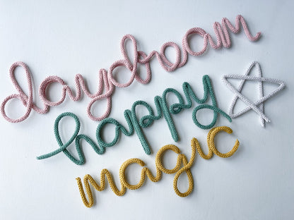 Flat lay style photo feature knitted wire words and wire art: daydream in the color blush, happy in the color sage green, magic in mustard and a whimsical star in light gray.