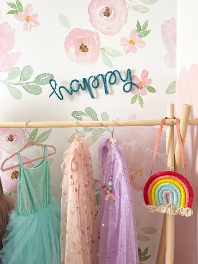 Knitted wire word "happy" featured in teal color hanging over dress up rack in kid's room.